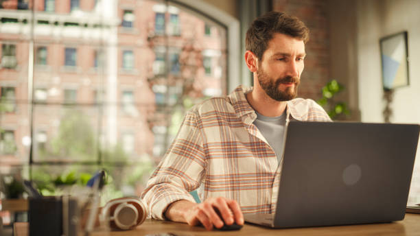 istock Portrait of a Handsome Man Working on Laptop Computer while Sitting in Stylish Cozy Loft Living Room. Freelancer Working From Home. Browsing Internet, Writing Emails, Being Productive 1478410820