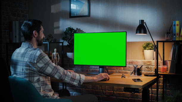 istock Young Attractive Man Working from Home on Desktop Computer with Green Screen Mock Up Display. Male Checking Corporate Accounts, Messaging Colleagues. Loft Living Room in the Evening 1478411464
