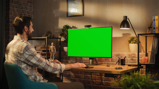 istock Young Professional Creative Specialist Works on His Personal Desktop Computer with Big Monitor with Green Screen Mock Up Display. He Works in a Cozy Office Loft in the Evening. 1478411647