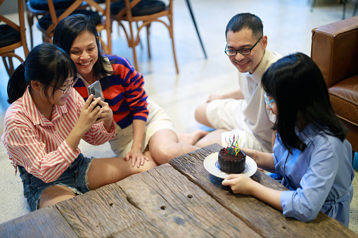 An Asian family celebrating the birthday of their youngest sister at home with a cake, using technology to capture the moment and promoting the values of bonding and lifestyle