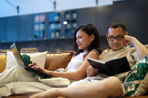 An Asian woman is using a laptop at home while her husband reads a book. They are both sitting on a comfortable leather sofa in the living room, spending quality time together.