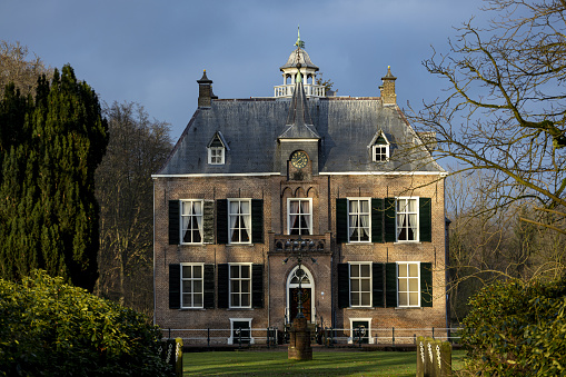 A large English Tudor Farm house with a gravel drive grass lawn and trees. The house has a red tiled roof and the scene is a summers day with blue sky and white clouds