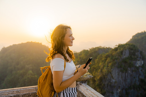 Young Caucasian woman  standing on the top of wat Tham Sua in Krabi at sunset and using smartphone