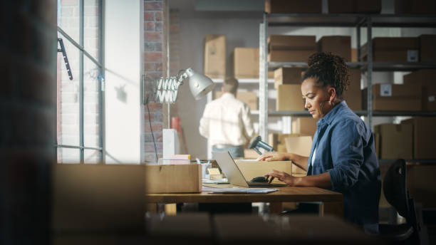 istock Black Female Warehouse Inventory Manager Works on Laptop Computer while Sitting at Her Desk. In the Background Employee Working in the Room Full of Cardboard Box Parcels Ready For Shipping. 1478421129