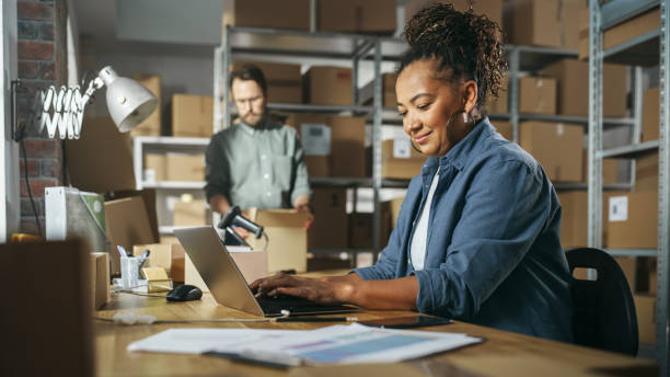 diversos gerentes de inventario de almacén masculinos y femeninos hablando, usando computadoras portátiles y verificando el stock minorista. filas de estantes llenos de paquetes de cajas de cartón en el fondo. - comercio electrónico fotografías e imágenes de stock