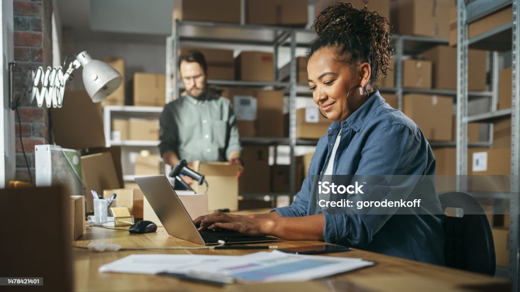 Diverse Male and Female Warehouse Inventory Managers Talking, Using Laptop Computer and Checking Retail Stock. Rows of Shelves Full of Cardboard Box Packages in the Background. Small Business Stock Photo
