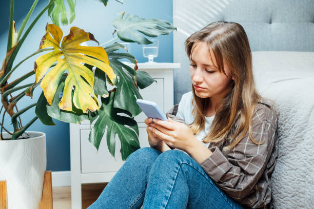istock Young woman using mobile phone for searching info about houseplants disorders, it's identification and treatment for dried, sunburn leaf of potted Monstera plant. Houseplants diseases. Home gardening 1478421530