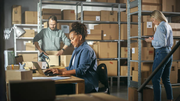 istock Establishing Shot of Multicultural Team of Warehouse Workers at Work in Internet Shop's Storeroom. Small Business Owners and Inventory Managers Working on Laptop, Tablet, Packing Parcels for Delivery. 1478421576