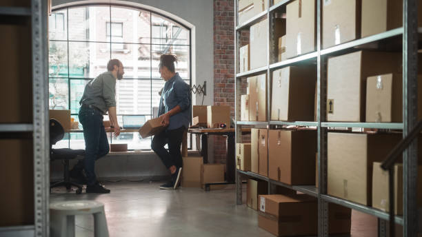 istock Establishing Shot of Warehouse Inventory Manager and Worker Using Laptop Computer, Having a Casual Chat. Small Business Owners Working in Storeroom, Preparing Online Orders for Customers. 1478421644
