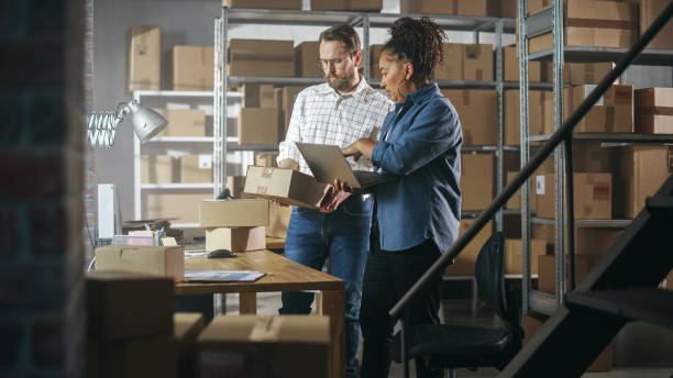 istock European Male and African Female Working in a Warehouse. Talking, Using Laptop Computer, Checking Retail Stock, Preparing Shipment. Rows of Shelves Full of Cardboard Box Parcels in the Background. 1478421709