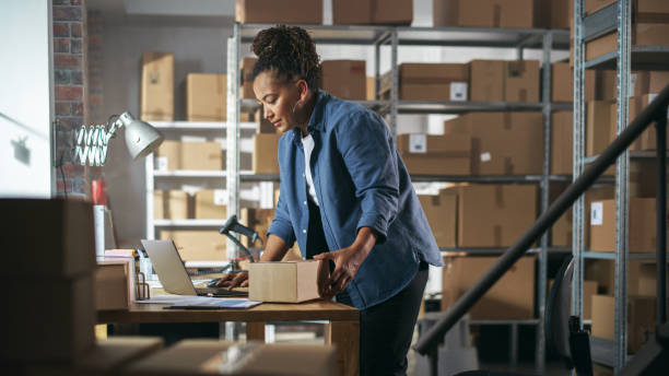 istock Female Inventory Manager Using Laptop Computer in Storeroom, Preparing a Small Cardboard Box for Postage. Black Multiethnic Small Business Owner Working in Warehouse, Fulfilling Order for Client. 1478422352