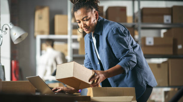 istock Inventory Manager Preparing a Small Cardboard Parcel for Postage. Multiethnic African American Female Small Business Owner Working on Laptop Computer in Warehouse 1478423567