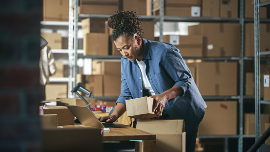 Inventory Manager Preparing a Small Cardboard Parcel for Postage. Multiethnic African American Female Small Business Owner Working on Laptop Computer in Warehouse.