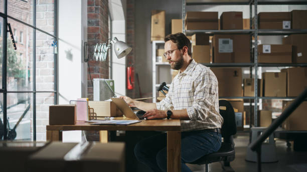 istock Inventory Manager Preparing a Small Cardboard Parcel for Postage. Stylish Young Male Small Business Owner Working on Laptop Computer in Warehouse Facility 1478423776
