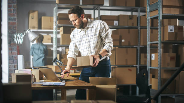 istock Inventory Manager Preparing a Small Cardboard Parcel for Postage. Stylish Young Male Small Business Owner Working on Laptop Computer in Warehouse Facility 1478423929