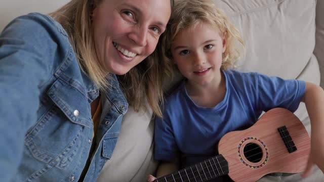 Mother making selfie video call whit child son playing ukulele inside living room at home - Family and technology concept