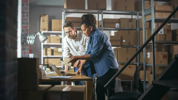 istock Multicultural Male and Female Warehouse Inventory Managers Talking, Using Laptop Computer and Checking Retail Stock. Rows of Shelves Full of Cardboard Box Packages in the Background. 1478424742