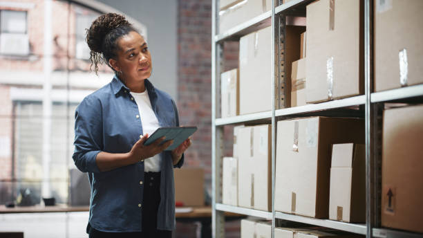 istock Portrait of a Worker Checking Inventory, Writing in Tablet Computer. Black Woman Working in a Warehouse Storeroom with Rows of Shelves Full of Parcels, Packages with Orders Ready for Shipment. 1478425672