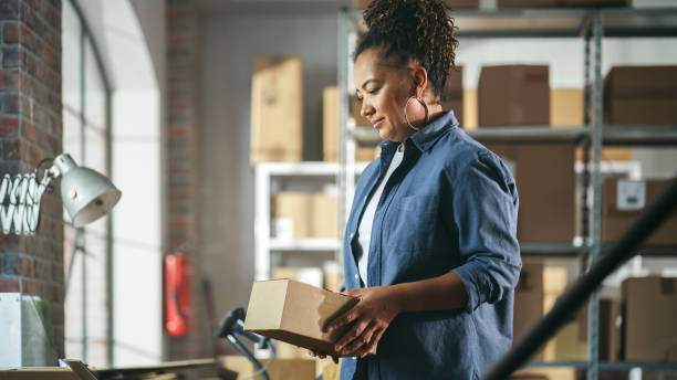 istock Small Business Owner Works on Laptop Computer while Standing at Her Desk in Warehouse. Employee Working in the Room with Shelves Full of Cardboard Box Parcels in the Background. 1478426195