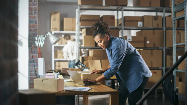 istock Warehouse Female Inventory Manager Works on Laptop Computer while Standing at Her Desk. In the Background Employee Working in the Room with Shelves Full of Cardboard Box Packages Ready For Shipping. 1478427066