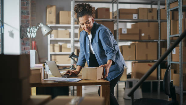 istock Warehouse Female Worker Using Laptop Computer, Preparing a Small Parcel for Shipping. African American Small Business Owner Working in Storeroom, Preparing Order for Client. 1478427120