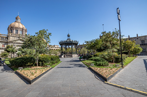 Panoramic view of the Plaza de Armas with a metal kiosk in background against blue sky, between cathedral and government building in historic city center, sunny day in Guadalajara, Jalisco Mexico