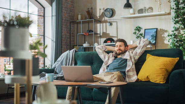 istock Happy Man Working from Home on Laptop Computer in Sunny Cozy Apartment, Relaxing on a Sofa. Successful Creative Male Entrepreneur Leans Back in Joy After Finishing Remote e Commerce Work 1478439916