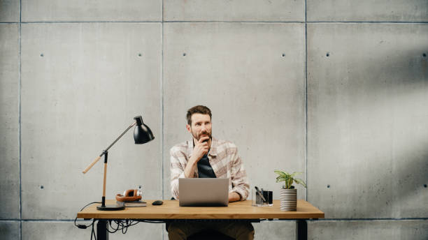 istock Professional Creative Man Sitting at His Desk in Office Working on a Laptop Computer, Working, Thinking, Solving Problems and Smiling at Success. Stylish Office Space Design Perfect for Productivity 1478441442