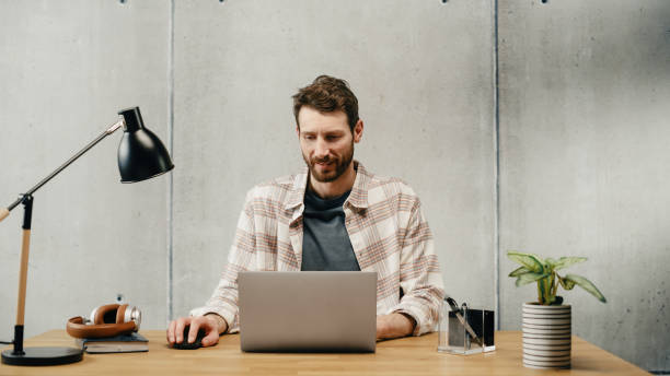 istock Professional Creative Man Sitting at His Desk in Office Working on a Laptop Computer. Stylish Clean Office Space Design Perfect for Productivity, Effectiveness and Vision. Medium Shot 1478441501