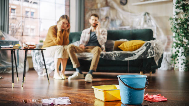 dach ist undicht oder rohrbruch zu hause: panikartiges paar in verzweiflung sitzt auf einem sofa und beobachtet, wie wasser in eimern in ihrem wohnzimmer tropft. katastrophe, unmut und finanzieller ruin - house repair stock-fotos und bilder