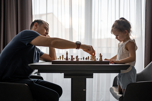 Little girl playing chess with her father at the table in home kitchen. The concept early childhood development and education. Family leisure, communication and recreation. High quality photo