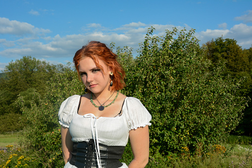 Portrait of a pretty young woman wearing white top, standing outdoors with green trees and blue sky