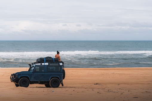 Newcastle, Australia – October 07, 2022: A Girl sitting on Land Rover 4WD with camping equiptment parked on stockton beach, Newcastle, NSW.