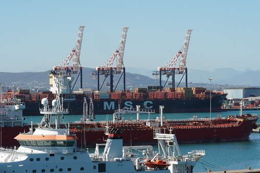 Cape Town, South Africa, 03 31 2023, Container ship from MSC company, orange tanker and white science vessel moored in port  Cape Town. In background are gantry crane and African mountains.