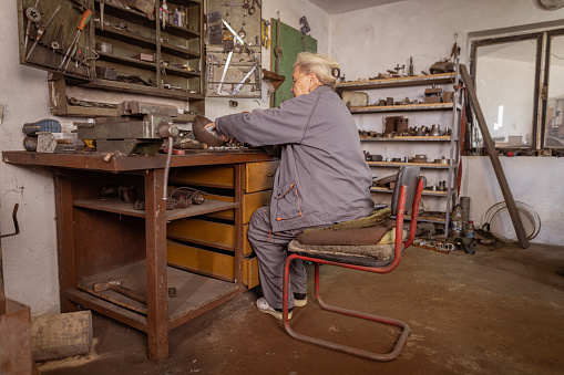 Elderly woman is sitting at the table in her workshop and working.