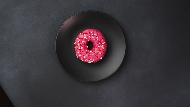 Top view of a Pink glazed Doughnut with colorful sprinkles on a black plate. Pink donut on a black background