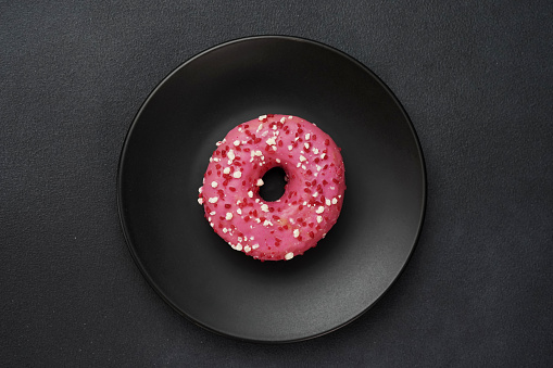 Top view of a Pink glazed Doughnut with colorful sprinkles on a black plate. Pink donut on a black background