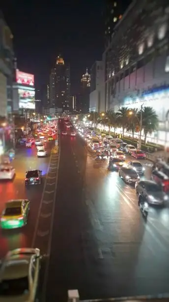 Experience the beauty of Bangkok's nightlife in the bustling city center with a view of a wide 3-lane street lined with towering buildings and advertising billboards. In the distance, you can see the silhouette of a towering skyscraper, while the busy traffic and glittering car lights create a mesmerizing scene.