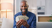 Smile, cash and investment with a black man in his home, sitting on the sofa holding dollar money as a lottery winner. Finance, economy or savings with a rich and happy young male in his living room