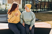 Two womans sitting outside on a bench and talking along on city street background. People and lifestyle concept