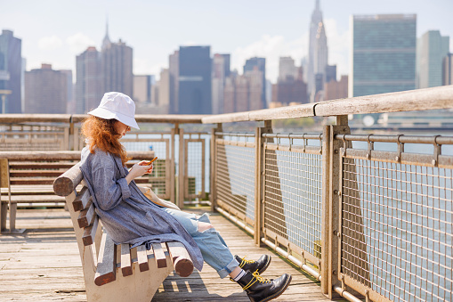 Young woman using her smartphone, sitting on the bench on a pier in Gantry Plaza State Park, Hunters Point, with Manhattan view behind.
