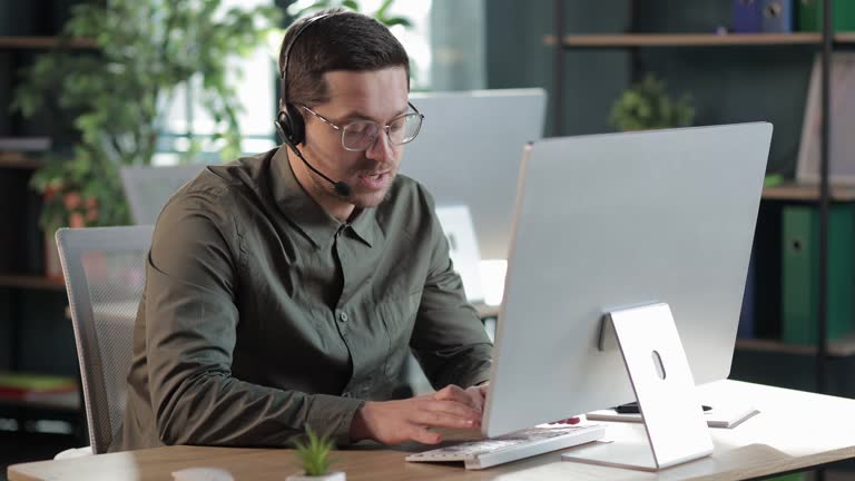 millennial man in glasses wearing headphones holding video call with clients partners. consulting online on helpline. talk with clients provide information strategizing remotely