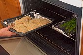 Placing roasting pan full of breaded chicken breasts into oven adjoining asparagus