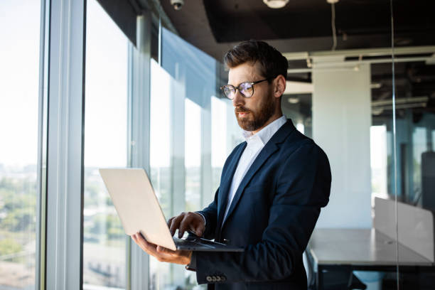 istock Successful middle aged businessman with laptop in hands standing in modern office interior near window, copy space 1478504368