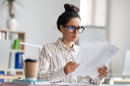 Paperwork concept. Focused female manager holding and reading paper or financial document, sitting at workplace in office. Woman checking reports