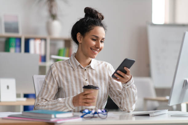 istock Happy female employee using smartphone and drinking coffee in office, having break at work, sitting at desk, free space 1478505221