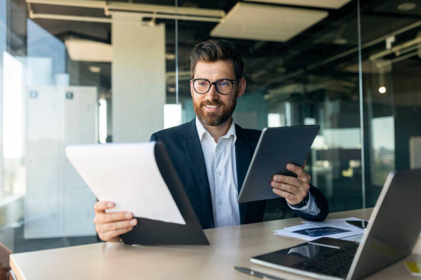 Cheerful middle aged businessman with documents and tablet working in office interior, free space Cheerful middle aged businessman with documents and tablet working in office interior, free space. Business, app for job, analyze finance, develop and startup general manager stock pictures, royalty-free photos & images