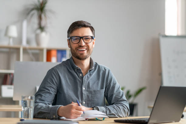istock Happy mature entrepreneur having online training in office, using laptop, taking notes and smiling at camera, copy space 1478505465