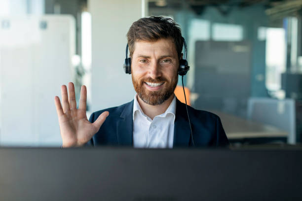ヘッドフォンを着た幸せな中年のビジネスマンがコンピューターのウェブカメラで手を振って、オフィスの内部で会議を開く - laptop men computer home interior ストックフォトと画像