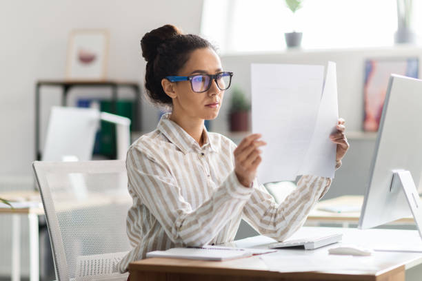 istock Serious busy woman in glasses sitting at table with computer and working with documents in office interior 1478505659